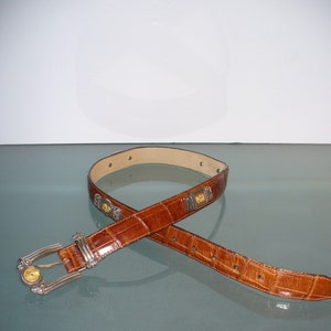 Pu&ograve; includere: Cintura in pelle marrone con motivo a coccodrillo. La cintura ha una fibbia argentata con dettagli dorati e abbellimenti decorativi argentati e dorati. La cintura &egrave; mostrata su una superficie riflettente.