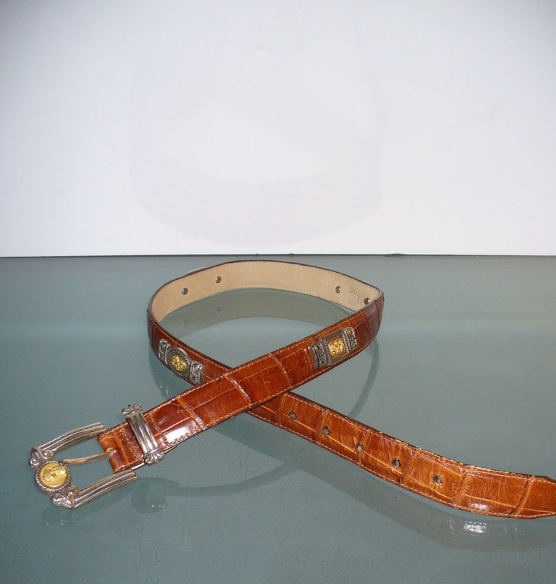 Pu&ograve; includere: Cintura in pelle marrone con motivo a coccodrillo. La cintura ha una fibbia argentata con dettagli dorati e abbellimenti decorativi argentati e dorati. La cintura &egrave; mostrata su una superficie riflettente.