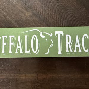 K&ouml;nnte beinhalten: Ein rechteckiger Aufkleber mit abgerundeten Ecken, mit den Worten "BUFFALO TRACE" in wei&szlig;er, stilisierter Schrift. Eine wei&szlig;e B&uuml;ffelkopf-Silhouette befindet sich auf der rechten Seite des Textes. Der Aufkleber hat einen hellgr&uuml;nen Hintergrund.