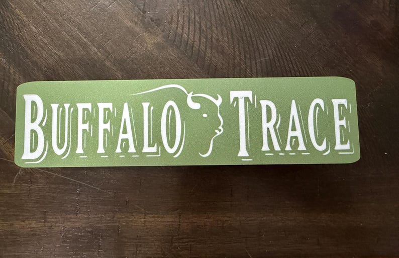 K&ouml;nnte beinhalten: Ein rechteckiger Aufkleber mit abgerundeten Ecken, mit den Worten "BUFFALO TRACE" in wei&szlig;er, stilisierter Schrift. Eine wei&szlig;e B&uuml;ffelkopf-Silhouette befindet sich auf der rechten Seite des Textes. Der Aufkleber hat einen hellgr&uuml;nen Hintergrund.
