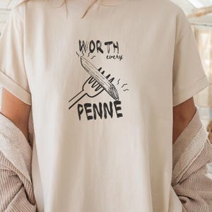 Peut inclure: T-shirt crème avec l'inscription noire "WORTH every PENNE", illustrée d'un dessin de pâtes penne sur une fourchette. Le t-shirt a des manches courtes et une coupe décontractée.