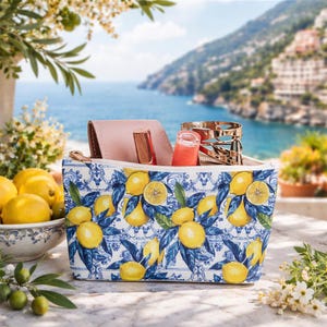 May include: A zippered cosmetic bag with a lemon and leaf pattern in yellow and blue. The bag contains a pink wallet, lipstick, and a gold bracelet. A bowl of lemons and a coastal scene are in the background.