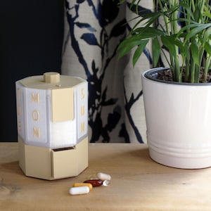 Op de afbeelding: Een beige, achthoekige pillendoos met een draaibaar bovendeel met doorschijnende panelen en gouden letters voor de dagen van de week, waaronder MON en SUN. De basis heeft een kleine lade en er liggen verschillende capsules voor het object.