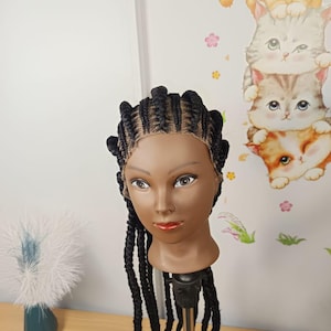 Peut inclure: Une tête de mannequin à la peau foncée avec de longs cheveux tressés noirs. Les tresses sont coiffées en épis sur le dessus, se transformant en longues tresses pendantes. Le mannequin a les yeux marron et du rouge à lèvres rouge. En arrière-plan, une décoration murale sur le thème des chats.