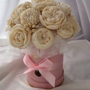 May include: A bouquet of ivory-colored flower-shaped candles arranged in a pink round box. The candles are designed to resemble roses, chrysanthemums, and other floral varieties. The box is adorned with a pink ribbon and a small decorative tag.