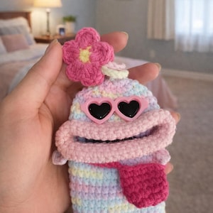 May include: A handmade crochet item in pastel colors, resembling a whimsical character. It features heart-shaped sunglasses, a protruding tongue, and a flower on top. The item is held in a hand.