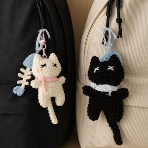 May include: Two crochet cat keychains attached to backpacks. One white cat with a pink scarf and a blue fish skeleton charm, and one black cat with a white scarf. Both cats feature embroidered X-shaped eyes and are suspended by metal clasps.