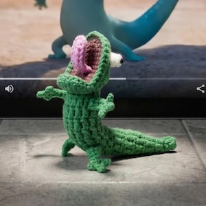 May include: A green crocheted alligator figurine with a pink mouth and white eye is in front of a laptop screen. The screen displays a cartoon lizard with its mouth open and tongue out. The background is a desert scene.