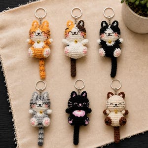 May include: Six crocheted cat keychains with silver rings. The cats feature various color patterns including orange tabby, calico, black and white, gray tabby, solid black with a pink chest patch, and Siamese-style, each with a small gold bell.