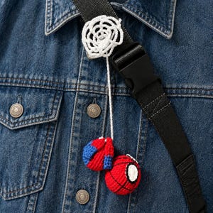 May include: A blue denim jacket with a black strap featuring a white crocheted spider web and two small, round, red and blue crocheted spheres with black web patterns attached by a thin white string, hanging against the denim fabric.