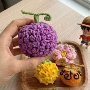May include: A collection of crocheted fruit-shaped items, including a purple, textured ball with a green stem, a pink flower, a yellow fruit, and an orange fruit with black swirls. A small figurine of a character wearing a straw hat is also present.