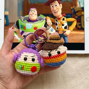 Puede incluir: Dos llaveros de ganchillo hechos a mano con personajes de la pel&iacute;cula de animaci&oacute;n "Toy Story". Un llavero representa a un extraterrestre morado y verde, y el otro a un personaje con un sombrero marr&oacute;n y una camisa a rayas de colores. Los llaveros se sostienen frente a una tableta que muestra una escena de la pel&iacute;cula.