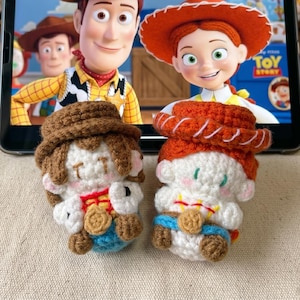 May include: Two crocheted Toy Story character dolls, Woody and Jessie, sit in front of a tablet displaying a scene from the movie. The dolls are white, brown, red, and blue, with detailed features. The background includes other Toy Story merchandise.