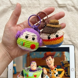 Puede incluir: Dos llaveros de ganchillo, uno morado y verde, el otro marr&oacute;n, naranja y azul. Los llaveros est&aacute;n dise&ntilde;ados para parecerse a personajes de la pel&iacute;cula de animaci&oacute;n "Toy Story". Los llaveros se sostienen frente a una tableta que muestra una escena de la pel&iacute;cula.