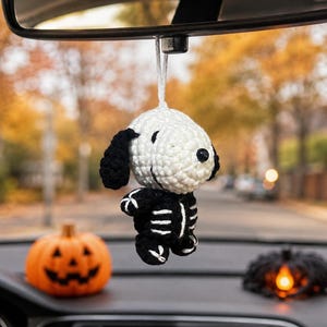 May include: A small, white and black crocheted dog figure wearing a black skeleton costume with white rib cage detailing. The figure is suspended by a white string from a rearview mirror, featuring a round head, floppy ears, and a black nose.