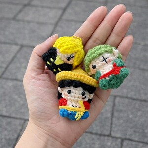 May include: Three small, handmade crochet character keychains. One has yellow hair and a black outfit, another has green hair and a red bow, and the third has a yellow hat and a red and blue outfit. The keychains are held in a hand.