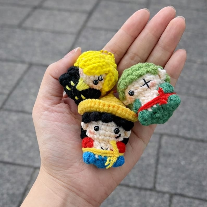 May include: Three small, handmade crochet character keychains. One has yellow hair and a black outfit, another has green hair and a red bow, and the third has a yellow hat and a red and blue outfit. The keychains are held in a hand.