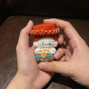 May include: A handmade crochet doll resembling a character from a popular animated film. The doll features a red hat, white face, and blue pants. It has a gold-colored medallion and brown boots. The doll is held in two hands.