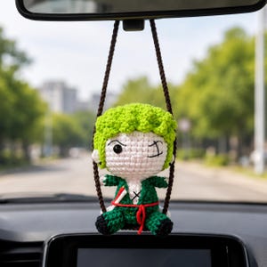 May include: A crocheted car charm featuring a character with green hair, a green outfit, and a red sash. The charm is suspended by brown cords and hangs from a car's rearview mirror. The background shows a blurred street scene with trees.