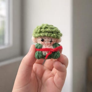 May include: A small, handmade crochet doll with a green cap, green legs, and a red sash. The doll has a light pink face with black eyes and a stitched cross on its face. The doll is held in a person's hand.