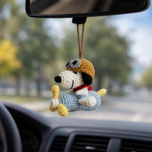 May include: Crocheted Snoopy car charm. The charm features Snoopy wearing a pilot's hat and goggles, seated in a blue airplane with yellow propellers. The charm is suspended from a brown string, designed to hang from a car's rearview mirror.