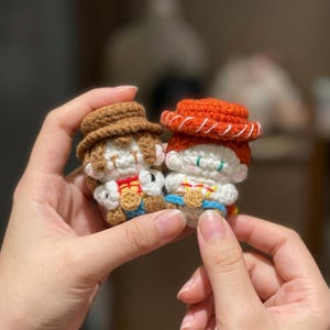 May include: Two small, crocheted figures of cartoon characters. One figure wears a brown hat and the other wears an orange hat. Both figures have white faces, and are wearing outfits with gold-colored badges. The figures are held in a person's hands.
