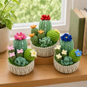 May include: Three crochet cactus arrangements in beige pots. The cacti are green with colorful flower accents in pink, orange, red, and blue. The arrangements sit on a wooden surface near a window.