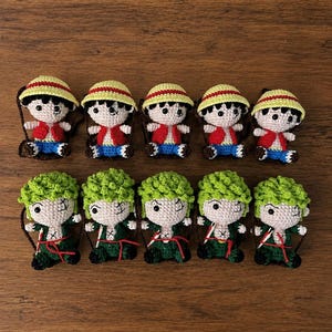 May include: Ten handmade crochet dolls. Five dolls are dressed in red vests, blue shorts, and yellow straw hats. The other five dolls have green hair and are dressed in green outfits. All dolls have black eyes and are on a brown background.