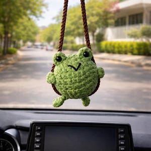 May include: A green crocheted frog car charm hanging from a brown cord. The frog has black eyes and a small smile. The charm is suspended in front of a car's dashboard, with a blurred street scene in the background.