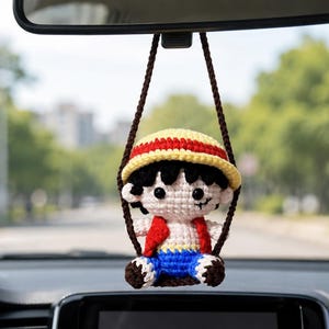 May include: Crocheted car charm featuring a cartoon character with a straw hat, red vest, blue shorts, and black hair. The charm is suspended from a brown string, hanging from a car's rearview mirror. The background shows a blurred street scene.