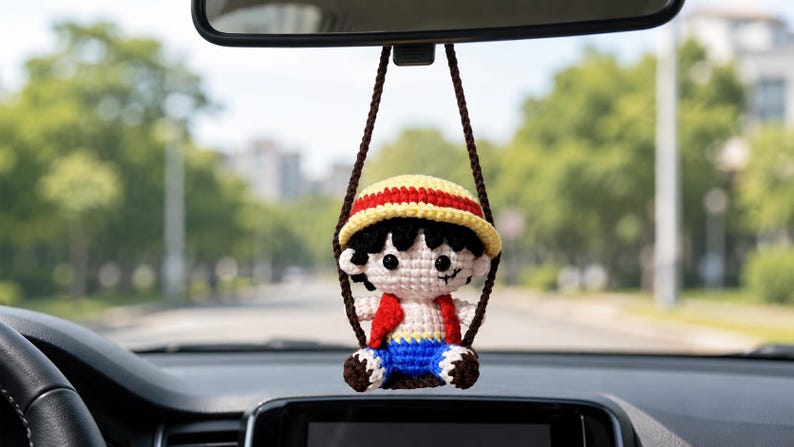 May include: Crocheted car charm featuring a cartoon character with a straw hat, red vest, blue shorts, and black hair. The charm is suspended from a brown string, hanging from a car's rearview mirror. The background shows a blurred street scene.