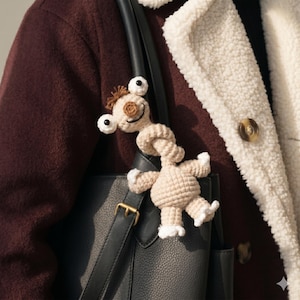 May include: A handmade, beige crochet creature with large, googly eyes and a smiling face. The whimsical figure has a long, twisted neck and small arms and legs with white accents. It is attached to a black leather bag.