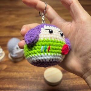 Puede incluir: Un llavero de ganchillo hecho a mano con un personaje redondo con orejas moradas, un cuerpo verde y blanco y una boca roja. El llavero tiene un anillo de metal plateado adjunto en la parte superior. La cara del personaje tiene ojos negros.