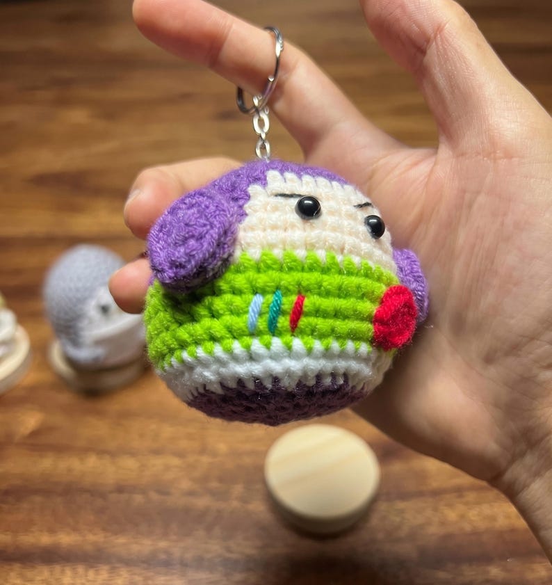 Puede incluir: Un llavero de ganchillo hecho a mano con un personaje redondo con orejas moradas, un cuerpo verde y blanco y una boca roja. El llavero tiene un anillo de metal plateado adjunto en la parte superior. La cara del personaje tiene ojos negros.