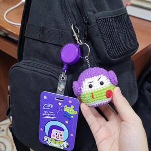 Puede incluir: Un portatarjetas retr&aacute;ctil morado con una tarjeta de identificaci&oacute;n de Buzz Lightyear y un llavero de personaje de ganchillo. La tarjeta de identificaci&oacute;n presenta a Buzz Lightyear y un gr&aacute;fico alien&iacute;gena con el texto "Oh! You got a friend!"