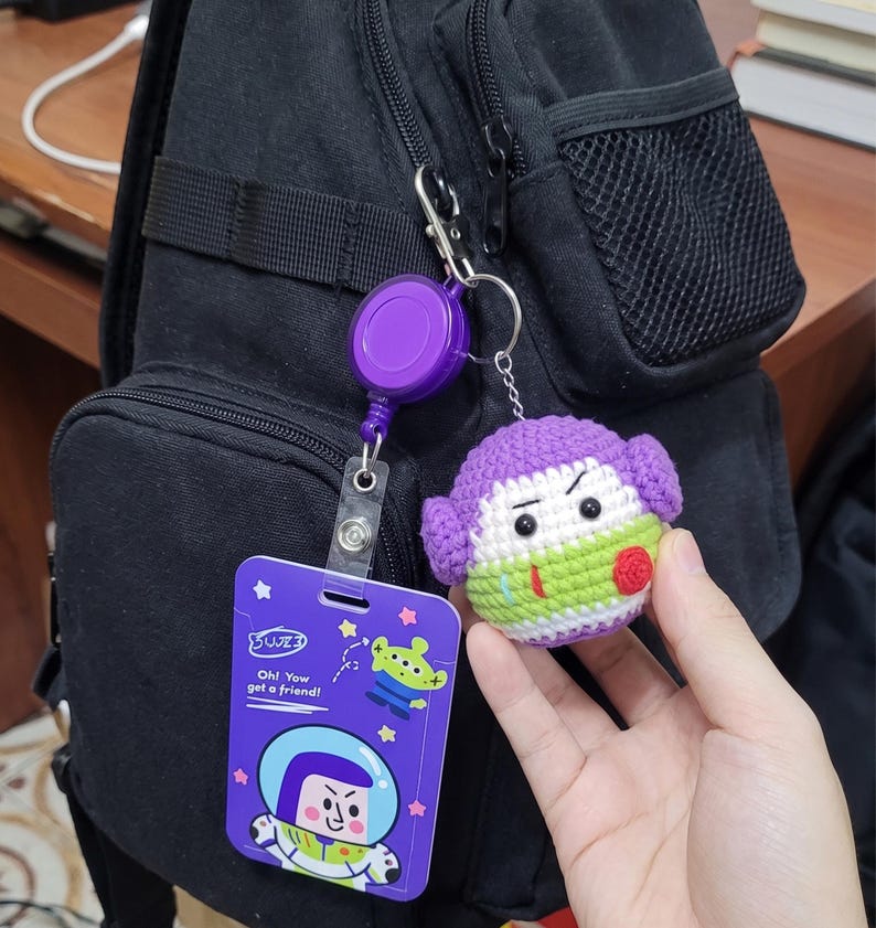 Puede incluir: Un portatarjetas retr&aacute;ctil morado con una tarjeta de identificaci&oacute;n de Buzz Lightyear y un llavero de personaje de ganchillo. La tarjeta de identificaci&oacute;n presenta a Buzz Lightyear y un gr&aacute;fico alien&iacute;gena con el texto "Oh! You got a friend!"