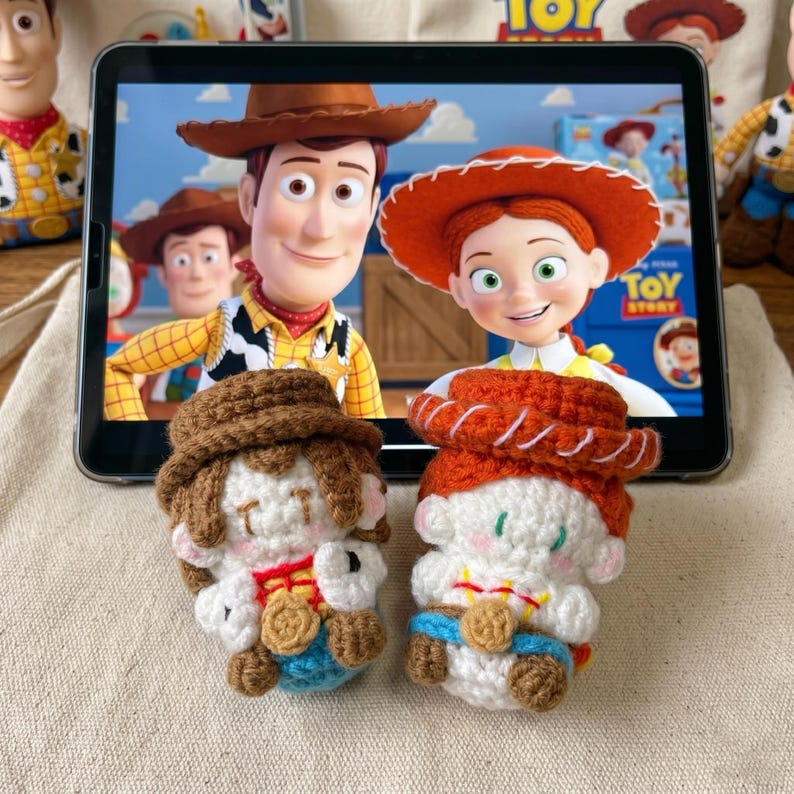 May include: Two crocheted Toy Story character dolls, Woody and Jessie, sit in front of a tablet displaying a scene from the movie. The dolls are white, brown, red, and blue, with detailed features. The background includes other Toy Story merchandise.