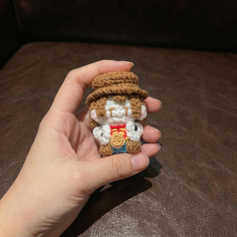 May include: A small, handmade crochet doll with a brown hat and hair, white face, and a red and gold outfit. The doll is held in a hand, showcasing the intricate details of the yarn work. The doll's design is reminiscent of a cartoon character.