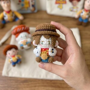 May include: A crocheted Woody doll from Toy Story, held in a hand. The doll has a brown hat, hair, and boots, with a yellow shirt, blue pants, and a red and yellow vest. Other crocheted dolls are in the background.