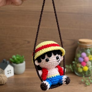 May include: A handmade crochet doll of a character wearing a yellow and red hat, red vest, and blue shorts, suspended from a brown string. The doll has black hair and is smiling. The background is a wooden surface.