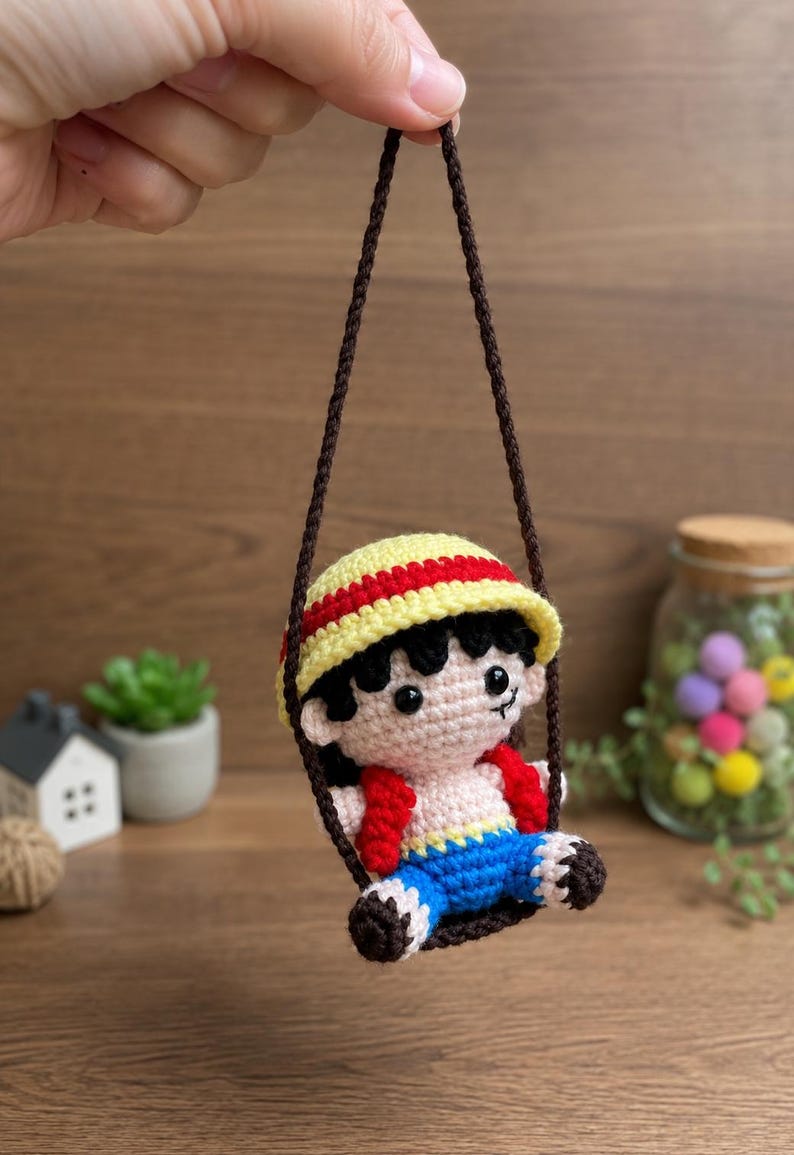 May include: A handmade crochet doll of a character wearing a yellow and red hat, red vest, and blue shorts, suspended from a brown string. The doll has black hair and is smiling. The background is a wooden surface.