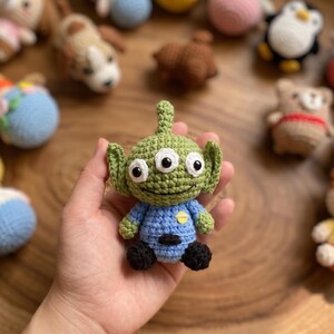 Puede incluir: Un juguete alien&iacute;gena verde tejido a ganchillo con tres ojos blancos, una camisa azul y pies negros. El alien&iacute;gena hecho a mano tiene orejas grandes y una cara sonriente. Otros juguetes tejidos a ganchillo est&aacute;n en el fondo.