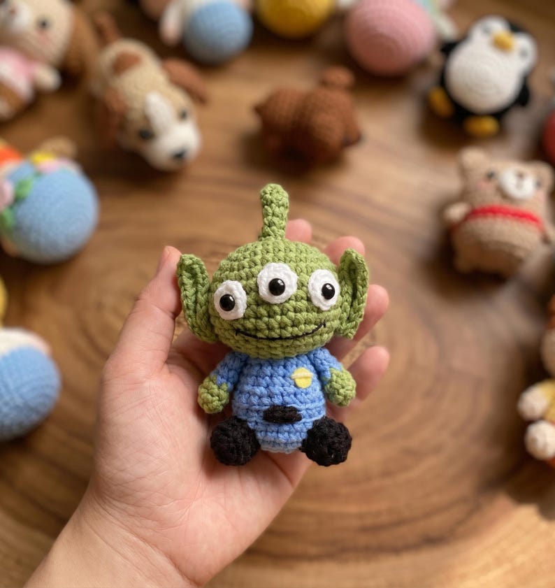 Puede incluir: Un juguete alien&iacute;gena verde tejido a ganchillo con tres ojos blancos, una camisa azul y pies negros. El alien&iacute;gena hecho a mano tiene orejas grandes y una cara sonriente. Otros juguetes tejidos a ganchillo est&aacute;n en el fondo.