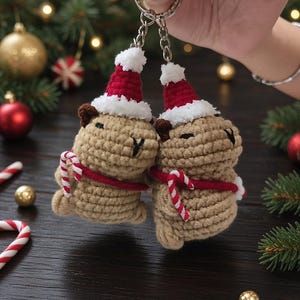 May include: Two crocheted capybara keychains made of tan yarn, each wearing a red and white Santa hat and a red scarf. Each capybara holds a small red and white striped candy cane. The figures are attached to a silver metal keyring and chain.