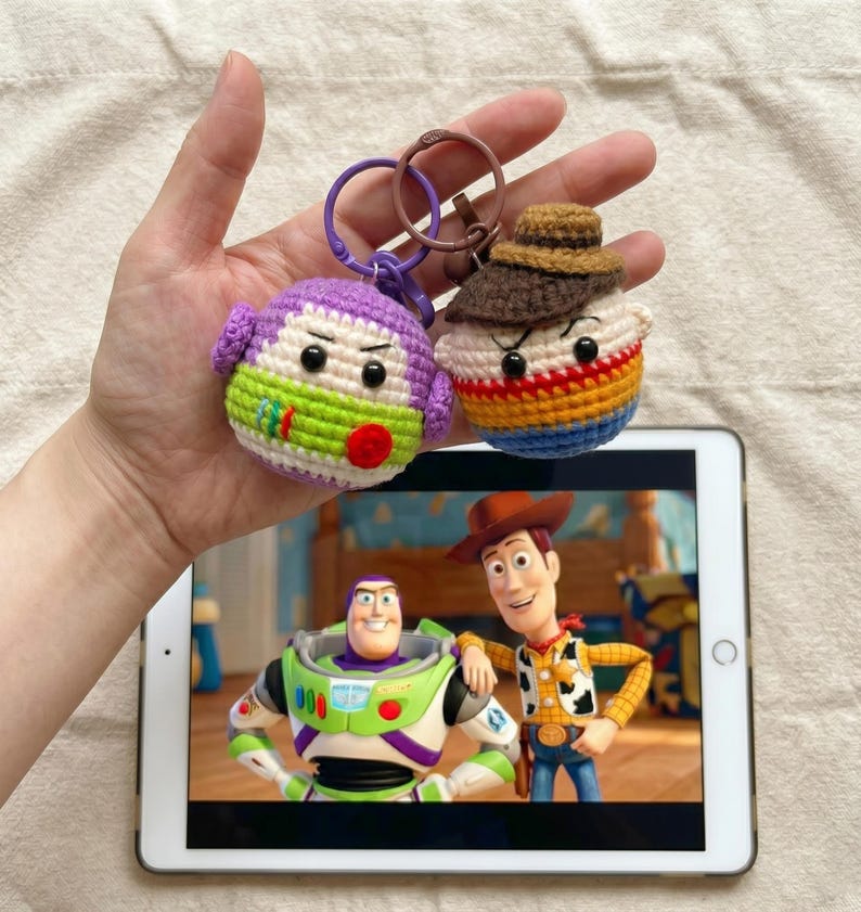 Puede incluir: Dos llaveros de ganchillo, uno morado y verde, el otro marr&oacute;n, naranja y azul. Los llaveros est&aacute;n dise&ntilde;ados para parecerse a personajes de la pel&iacute;cula de animaci&oacute;n "Toy Story". Los llaveros se sostienen frente a una tableta que muestra una escena de la pel&iacute;cula.