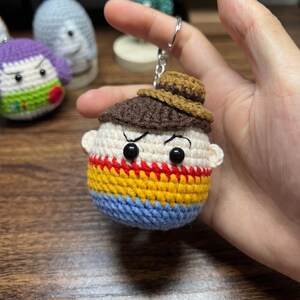 Puede incluir: Un llavero de ganchillo hecho a mano que representa un personaje de dibujos animados con un sombrero marr&oacute;n y un cuerpo a rayas en azul, amarillo y rojo. El llavero tiene un anillo de metal plateado. El personaje tiene ojos negros y una expresi&oacute;n severa.