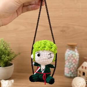 May include: A handmade crochet doll of a character with green hair and a green outfit, winking. The doll is suspended by a brown string. The doll is approximately 4 inches tall.