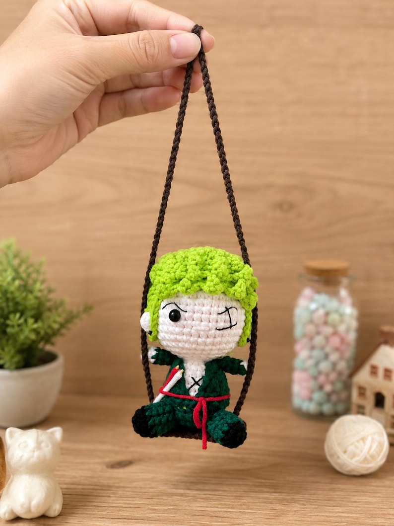 May include: A handmade crochet doll of a character with green hair and a green outfit, winking. The doll is suspended by a brown string. The doll is approximately 4 inches tall.