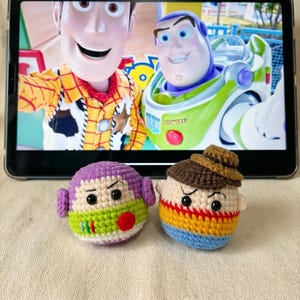Puede incluir: Dos mu&ntilde;ecos amigurumi de ganchillo de los personajes de Toy Story, Buzz Lightyear y Woody, est&aacute;n sentados frente a una tableta que muestra una escena de la pel&iacute;cula. El mu&ntilde;eco de Buzz es morado, verde y rojo. El mu&ntilde;eco de Woody es marr&oacute;n, amarillo y azul.