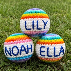 May include: Three crocheted balls with rainbow stripes and white bases, each embroidered with a name in blue: Lily, Noah, and Ella. The balls are arranged on a bed of green grass.
